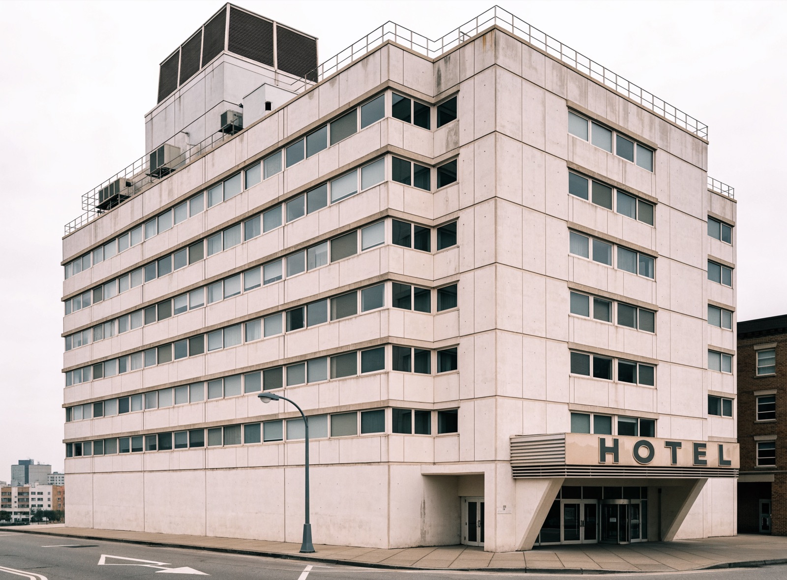 An operating mid-century full-service hotel in precast concrete, with a rooftop cooling tower, staining below the parapet and original ribbon glazing.